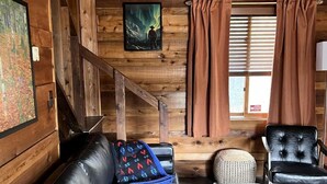 Ein Interieur einer Blockhütte mit einem schwarzen Ledersofa, einer grünen Bank, einem schwarzen Leder-Sessel und einer Treppe.