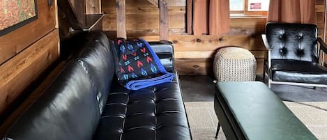 Interior de una cabaña de madera con un sofá de cuero negro, un banco verde, un sillón de cuero negro y una escalera.