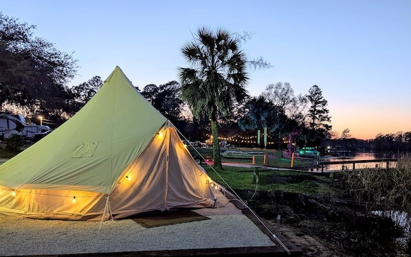 A large, light-colored tent with string lights, set up on a gravel area near a body of water, with trees and a bridge in the background.