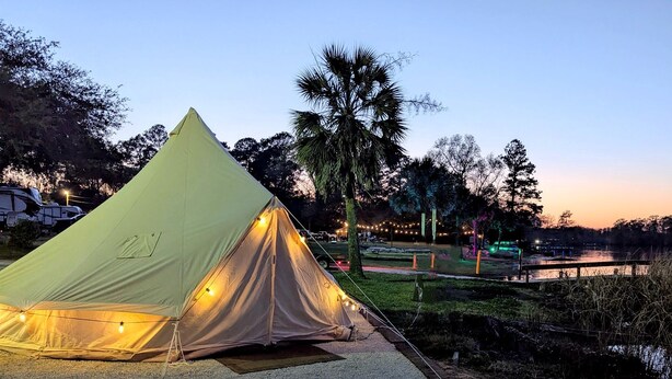 Una carpa grande de color claro con luces de cadena, montada sobre una zona de grava cerca de un cuerpo de agua, con árboles y un puente al fondo.