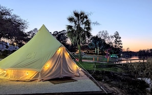 A large, light-colored tent with string lights, set up on a gravel area near a body of water, with trees and a bridge in the background.