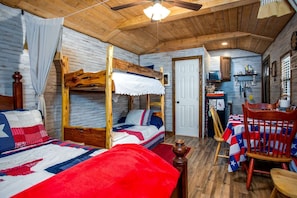 Ein rustikaler Raum im Blockhausstil mit einem Etagenbett, einem Esstisch mit rot-weißer Tischdecke und einem hölzernen Deckenventilator.