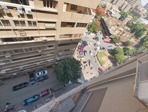En hög byggnad med flera balkonger, en parkeringsplats med bilar och en utsikt över gatan med träd och andra byggnader.