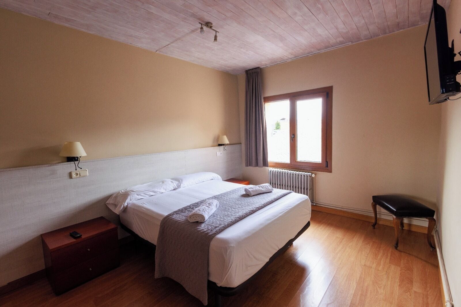 A bedroom with a bed, bedside lamps, a wooden nightstand, a radiator, and a window with curtains.
