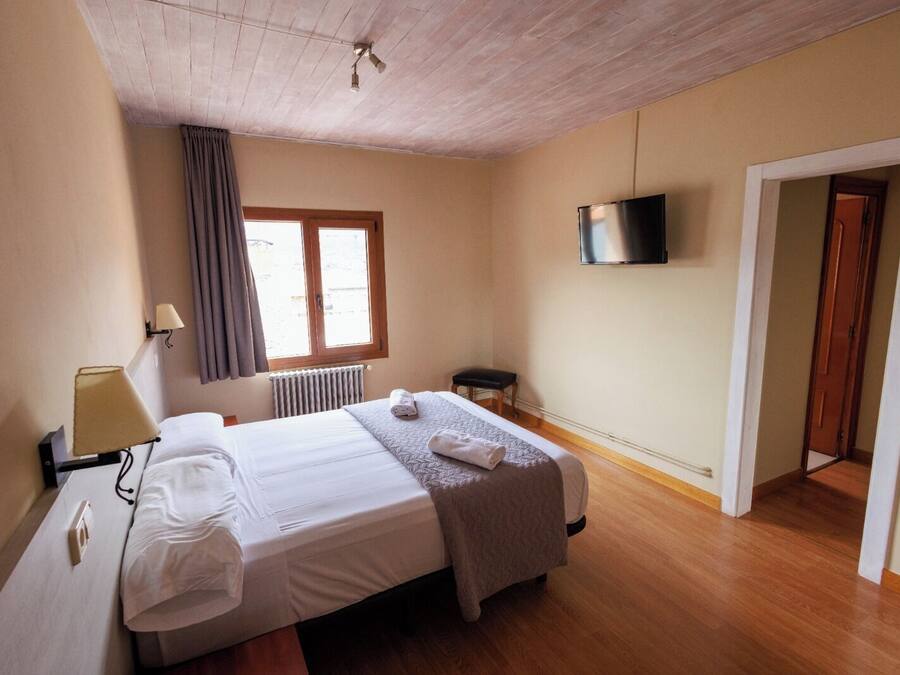 Habitación de hotel con cama, escritorio, silla, radiador, ventana con cortinas, televisor y puerta.