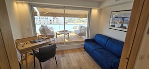 A compact living space with a wooden desk, a blue sofa, and a view of a pool and buildings. - Top floor apartment with panoramic view (Antibes)