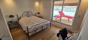 Ein Schlafzimmer mit Bett, Nachttischen, einem Fenster mit Blick auf einen Balkon und einer Tür.