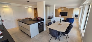 A modern kitchen with a central island, a dining area with a wooden table and chairs, and a living space with a TV and wooden paneling. - Top floor apartment with panoramic view (Antibes)