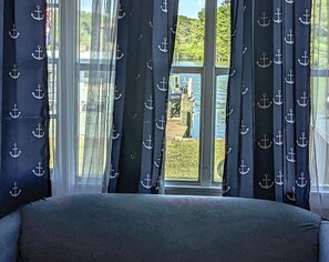Une chambre avec une grande fenêtre, des rideaux ornés de motifs d’ancres et une vue sur un quai et la verdure environnante.