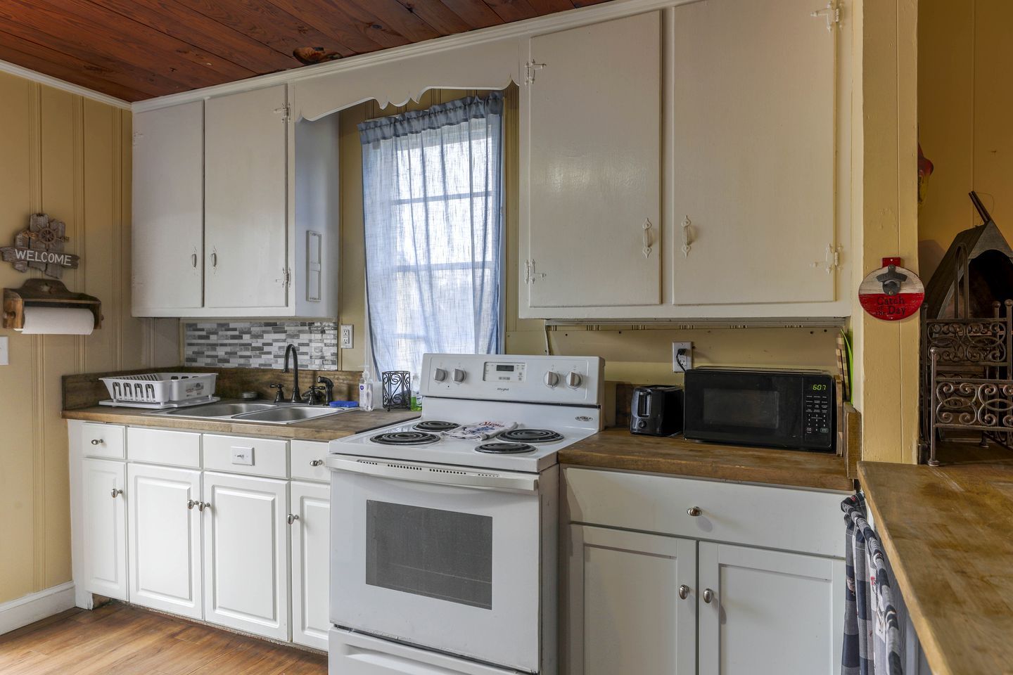 A kitchen with white cabinets, a stove, a microwave, and a window with a curtain.