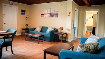 A living room with a blue sofa, wooden coffee table, and a dining area with a table and chairs. An exit sign is visible on the wall.