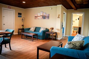 Un salon avec un canapé bleu, une table basse en bois et un coin repas comprenant une table et des chaises. Un panneau indiquant la sortie est visible sur le mur.