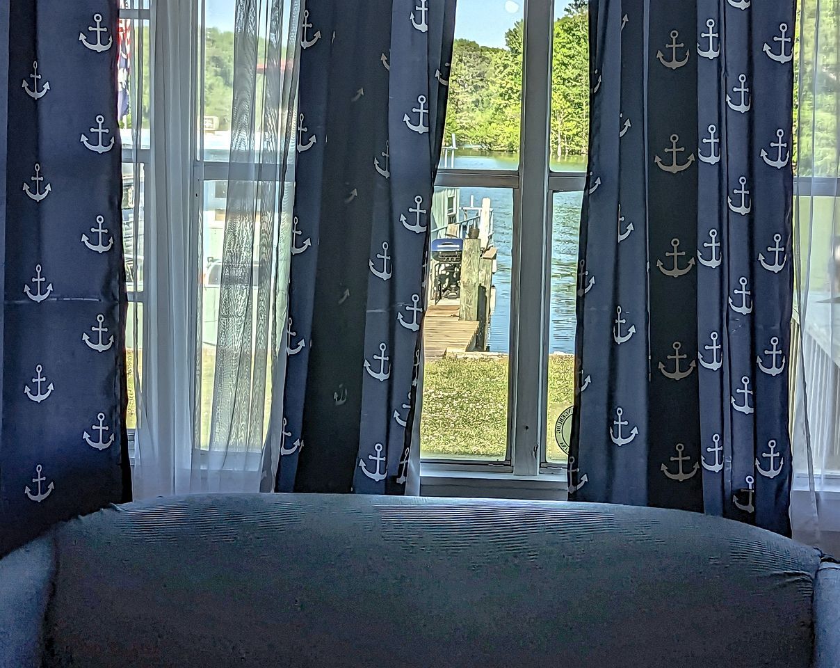 A bedroom with a large window, curtains with anchor patterns, and a view of a dock and greenery outside.