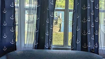 A bedroom with a large window, curtains with anchor patterns, and a view of a dock and greenery outside.