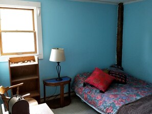 Ein Zimmer mit blauer Wand, einem Bett mit floralem Bettbezug, einem roten Kissen, einer Lampe auf einem Nachttisch und einem Holzstuhl.
