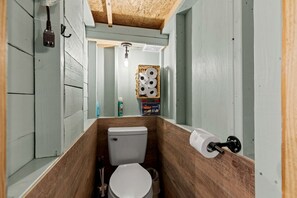 Une petite salle de bain avec un plancher en bois, des toilettes blanches, un luminaire suspendu et un panier rempli de papier toilette.