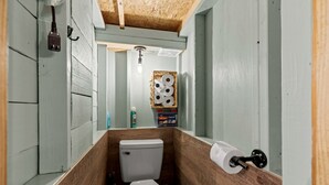 A narrow bathroom with a wooden floor, a white toilet, a hanging light fixture, and a basket of toilet paper.
