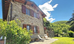 Un bâtiment en pierre avec des volets en bois, une terrasse avec des chaises et un jardin luxuriant.