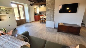 Ein Wohnzimmer mit einer Couch, einem an einem Steinsäule befestigten Fernseher und einer Küche im Hintergrund.