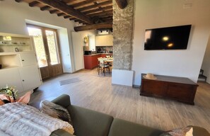 Un salon avec un canapé, un téléviseur fixé sur un pilier en pierre, et une cuisine en arrière-plan.
