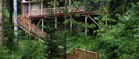 Una cabaña con terraza de madera y escalera, rodeada de un bosque denso.
