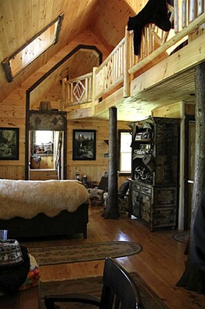 A wooden cabin interior with a loft bedroom, a dining area, and a fireplace.