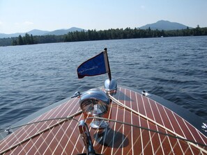 Cubierta de barco con una bandera, un timón y una vista de un lago tranquilo con montañas al fondo.