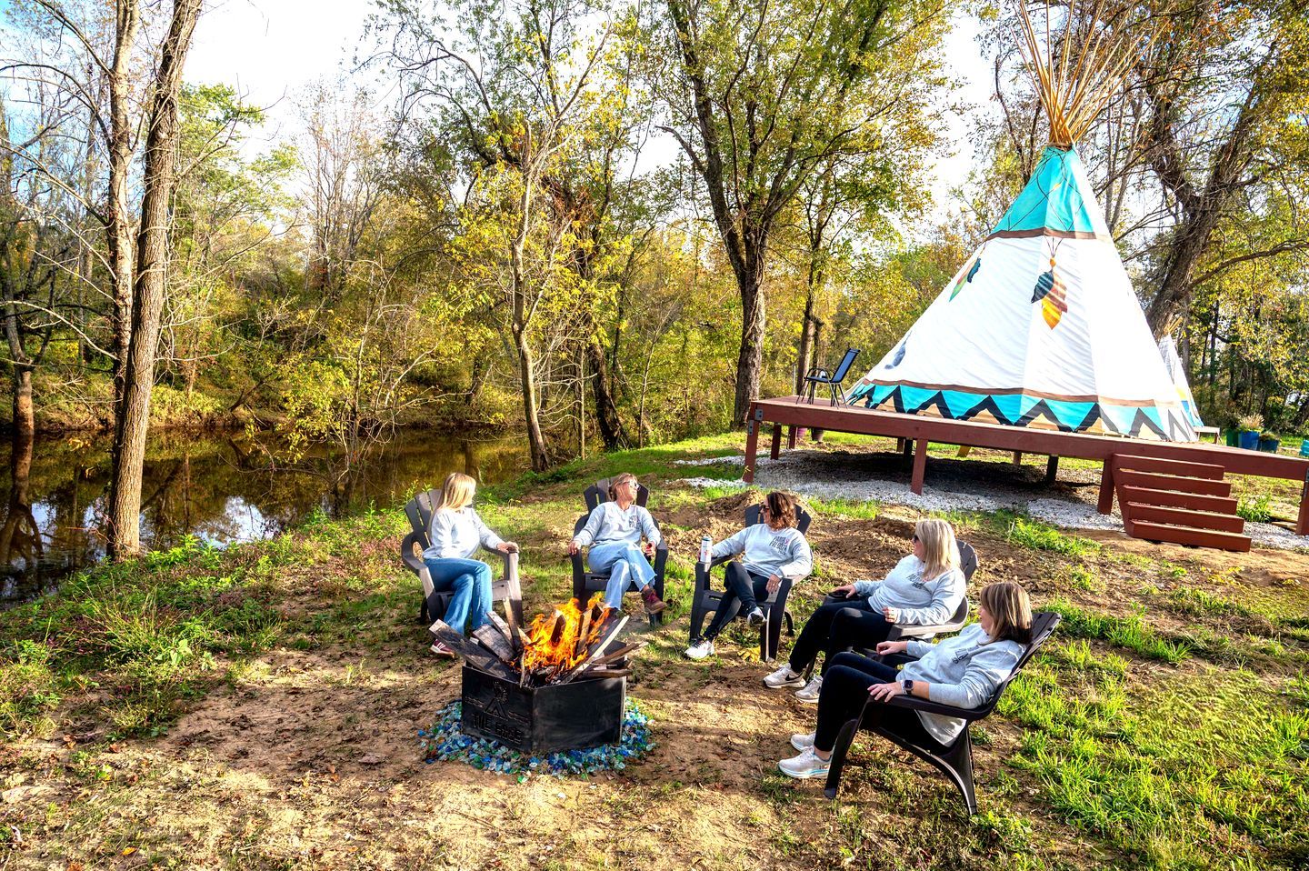 A group of people seated around a campfire in a forested area with a teepee structure in the background.