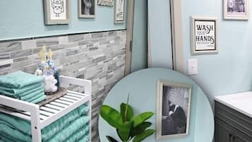 A bathroom with a white bathtub, a sink, and a toilet. There are towels stacked on a rack, a potted plant, and framed pictures on the wall.