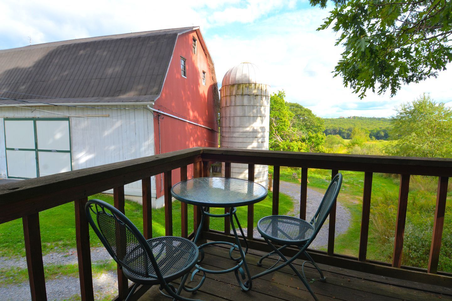 A wooden deck with a table and two chairs, overlooking a red barn and a silo.