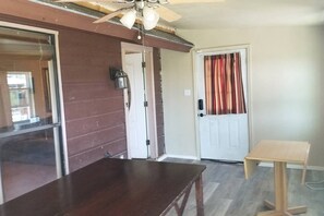 A room with a wooden table, a ceiling fan, a door with a curtain, and a small wooden stool.