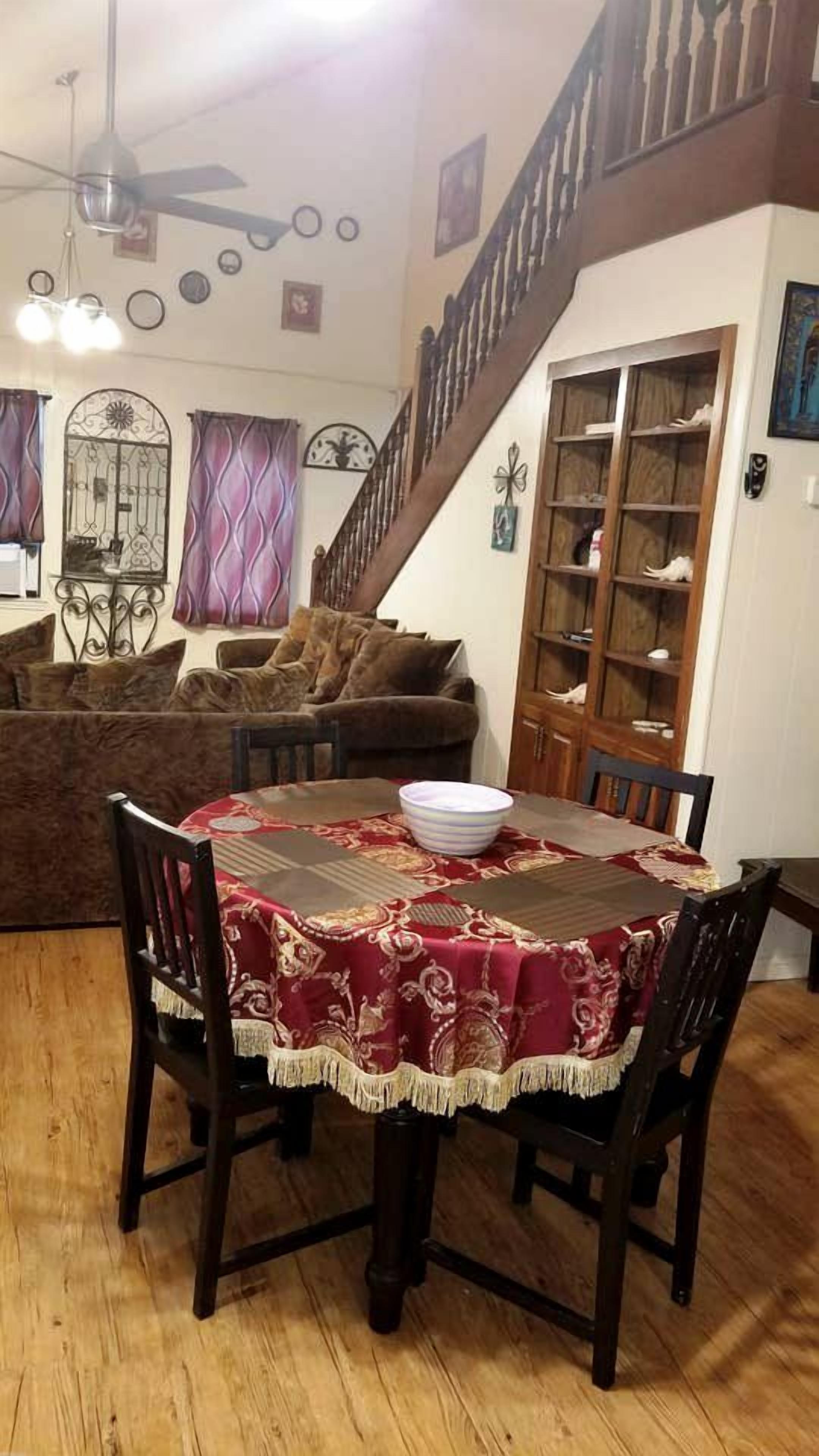 A dining area with a round table, chairs, and a red tablecloth. There is a staircase with a railing and a wooden cabinet with glass doors.