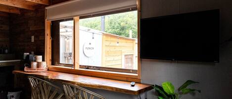 A room with a wooden desk, two chairs, a TV, and a window with a view of a wooden fence and greenery.