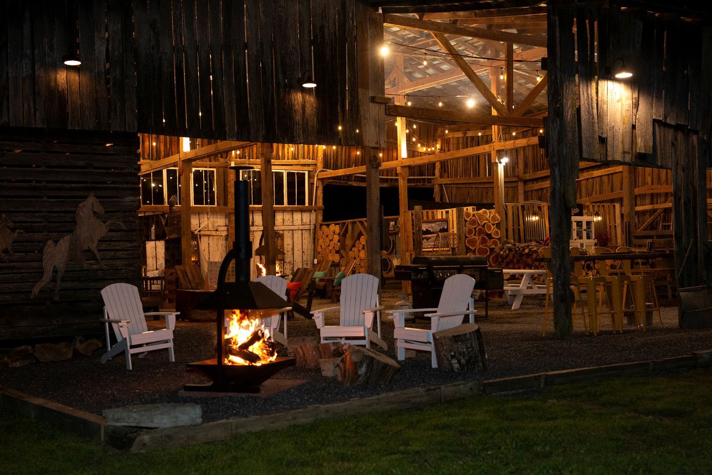 A rustic wooden building with a fire pit, white Adirondack chairs, and a dining area.