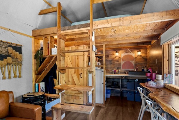 L’intérieur d’un chalet rustique comprend un escalier en bois, un coin cuisine et un espace salon avec une table et des chaises.