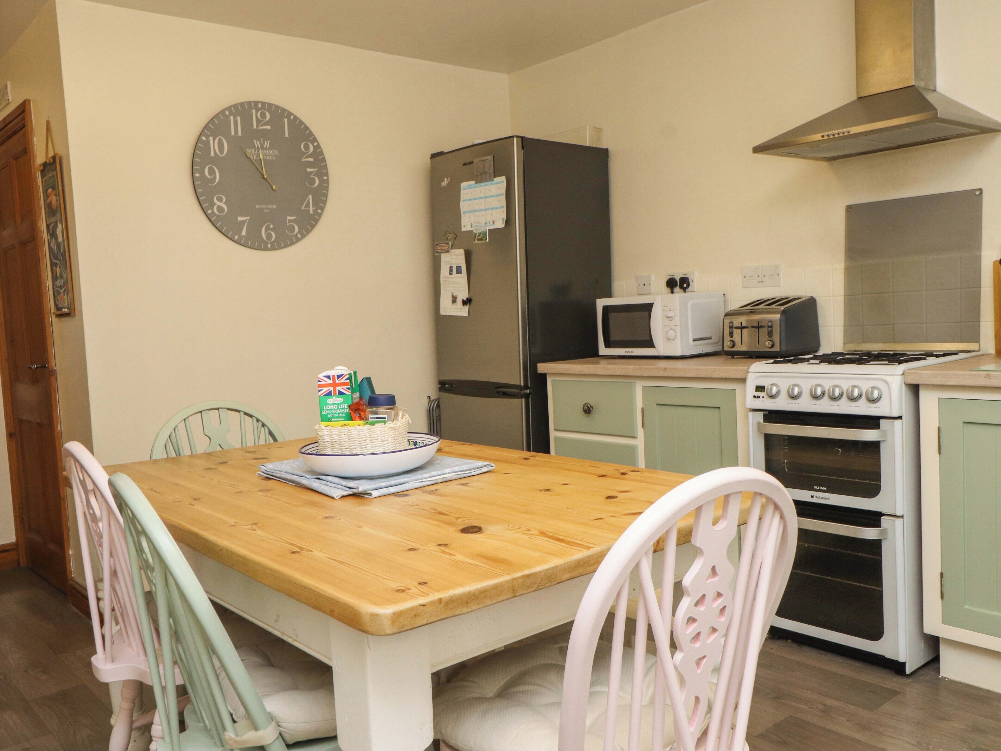 A kitchen with a wooden table, chairs, a clock, a refrigerator, a microwave, a toaster, and an oven.