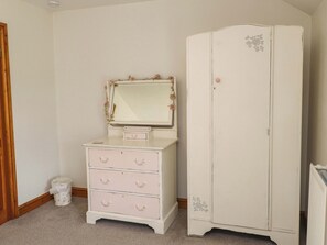 Una habitación con un aparador blanco con cajones y un espejo, un armario blanco con motivos florales y un pequeño cubo de basura.