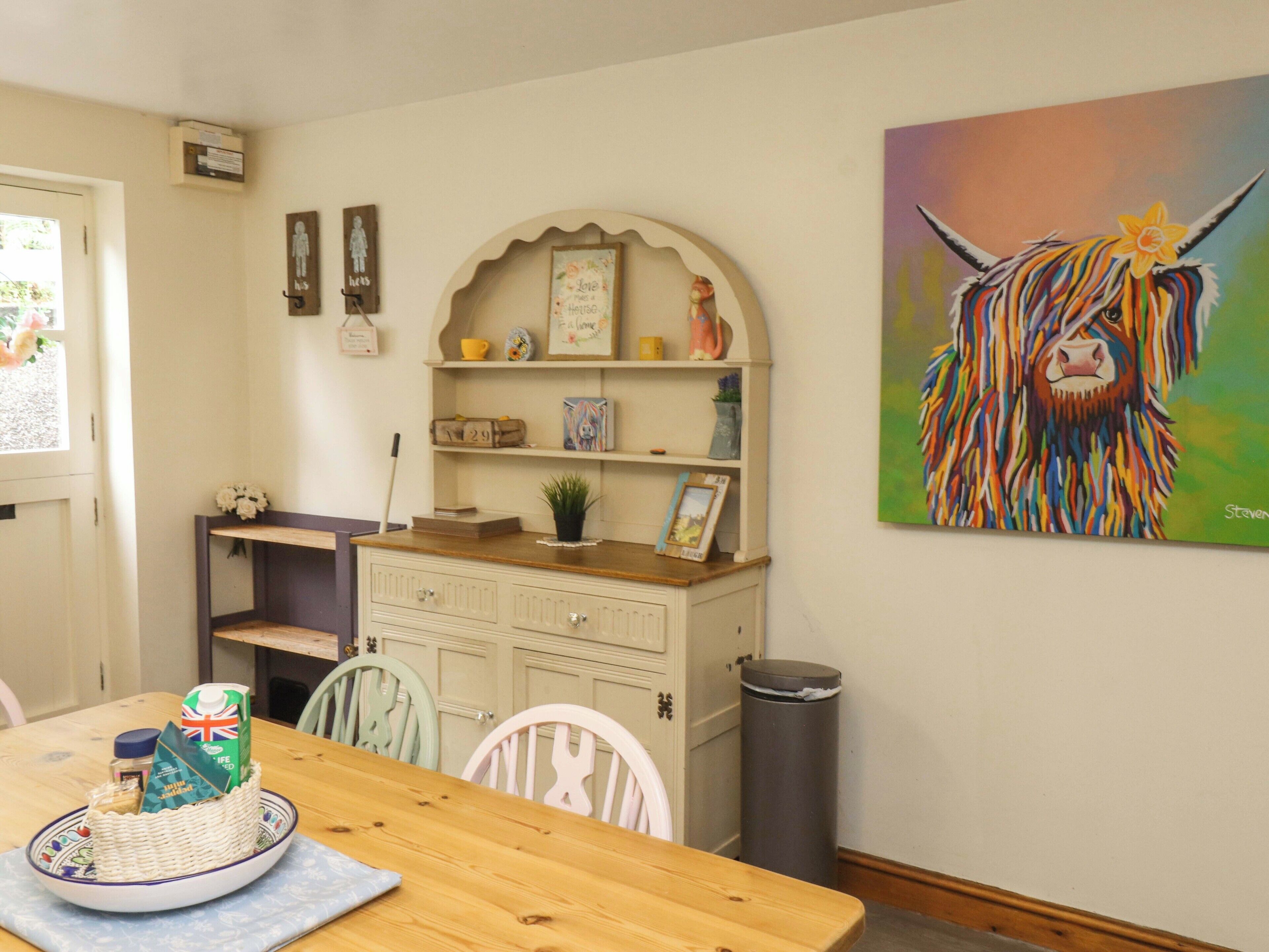 Un comedor con mesa de madera, sillas, una cesta de comida, estante con adornos y un cuadro de un animal colorido.