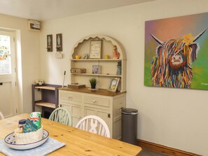 A dining area with a wooden table, chairs, a basket of food, a shelf with decorative items, and a painting of a colorful animal. - 2 Bryn Eglwys (Porthmadog)
