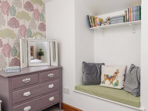 Quarto com cômoda, espelho, estante de livros e um banco com almofadas.