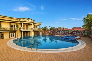 Uma grande piscina ao ar livre com deck de madeira, um edifício com sacadas e um muro colorido ao fundo.