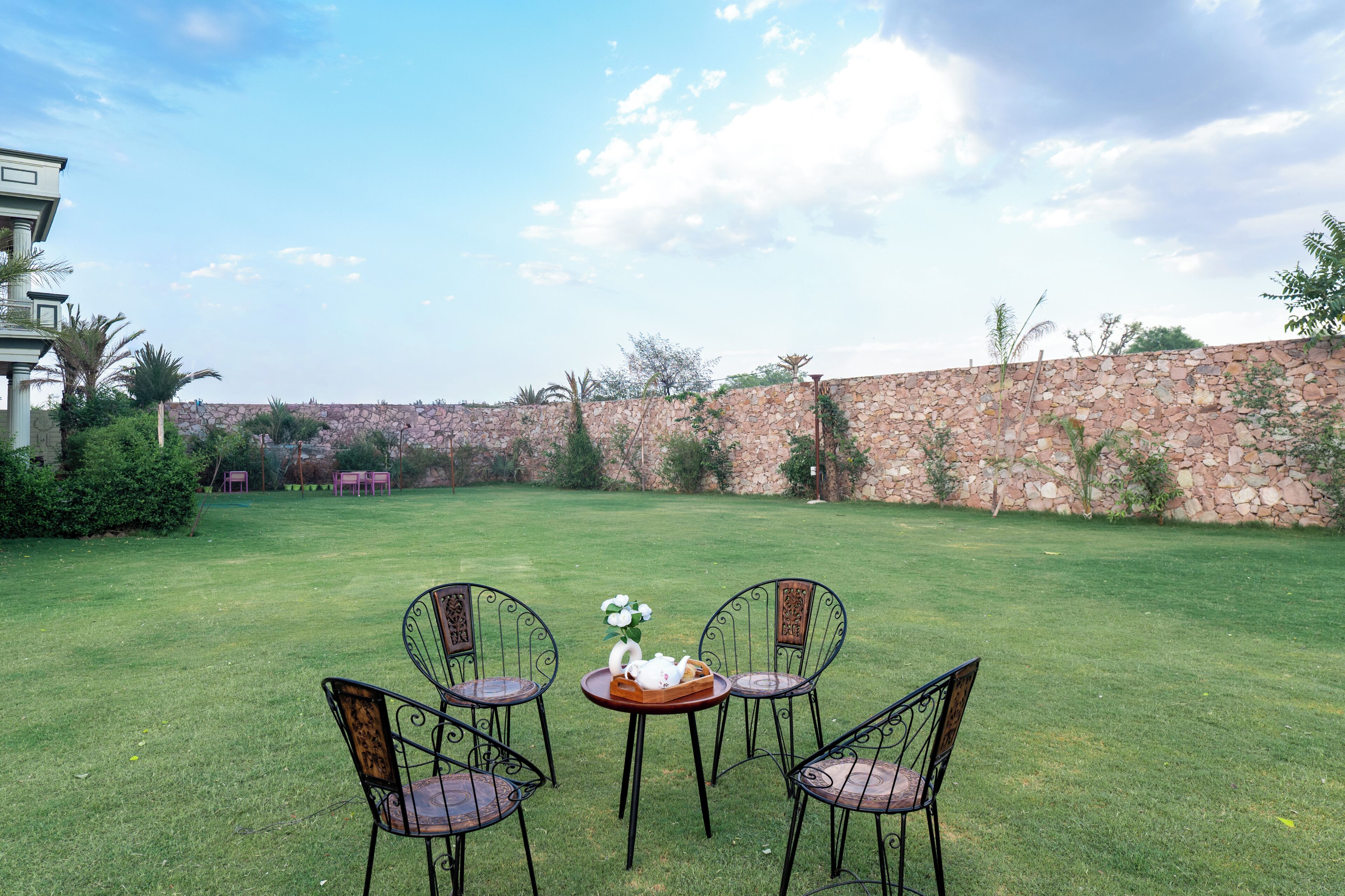 A grassy outdoor area with a stone wall, four metal chairs, and a table with a teapot and flowers.