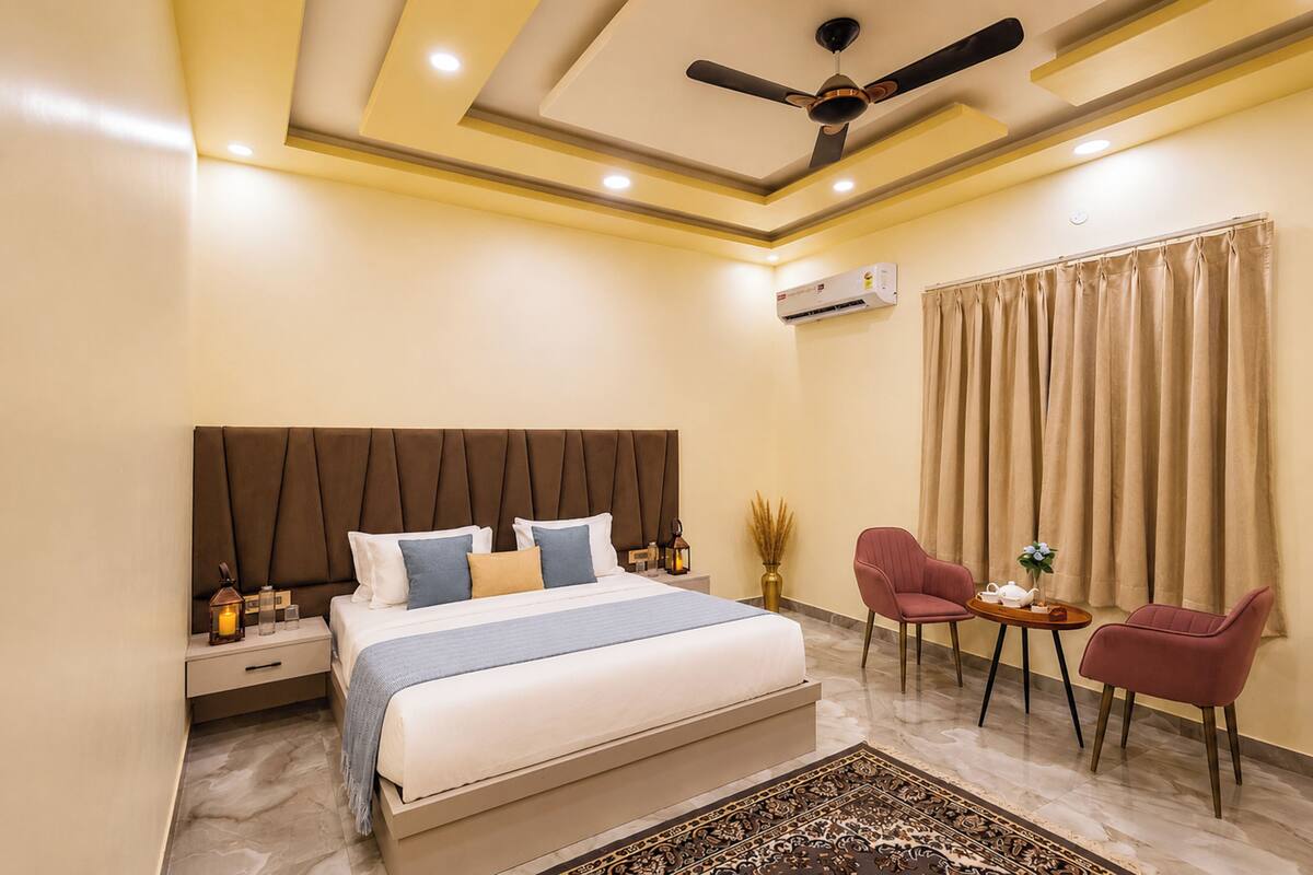 Un dormitorio con cama, mesitas de noche, ventilador de techo, aire acondicionado, ventana con cortinas y dos sillas.
