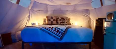 Una cama dentro de una tienda de campaña, con una colcha estampada, almohadas y una mesita con una lámpara.