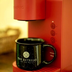 Uma máquina de café com exterior vermelho e uma caneca de café preta com a inscrição 'THE RETREAT ON THE MILE'.