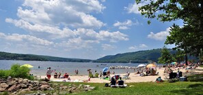 Una playa junto al lago con gente nadando y tomando el sol, rodeada de vegetación y colinas.