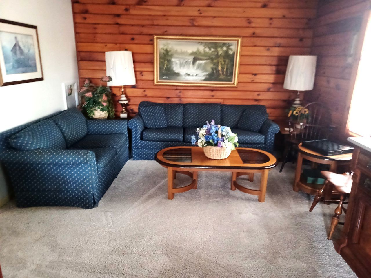 Un salon avec un canapé bleu à motifs, une table basse en bois agrémentée d’un arrangement floral, et deux lampes.