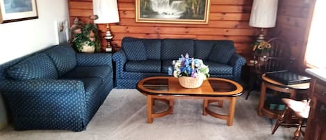 Uma sala de estar com um sofá azul estampado, uma mesa de centro de madeira com um arranjo floral e duas luminárias.