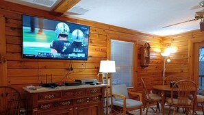 A wooden interior with a television displaying a sports game, a dining table with chairs, a lamp, and a bookshelf. - Family and Pet-Friendly Cabin Rental with a Dock Slip near the Wisp Resort, Maryland (Swanton)