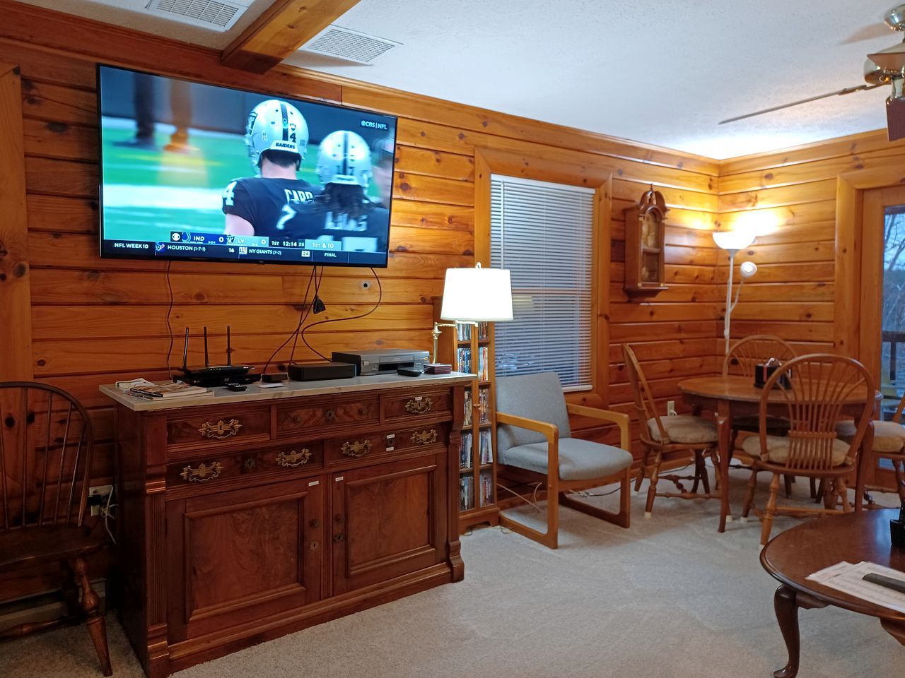 A wooden interior with a television displaying a sports game, a dining table with chairs, a lamp, and a bookshelf.
